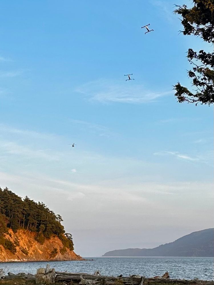 Picturesque view of the ocean and distant island at golden hour. In the sky there is a helicopter and 2 large passenger drones. 
