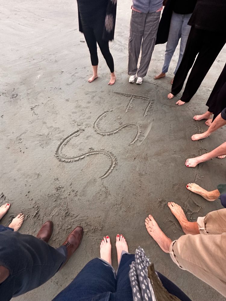 Photo shows 10 pairs of bare feet around the letters "FSS" written in sand on a beach. 