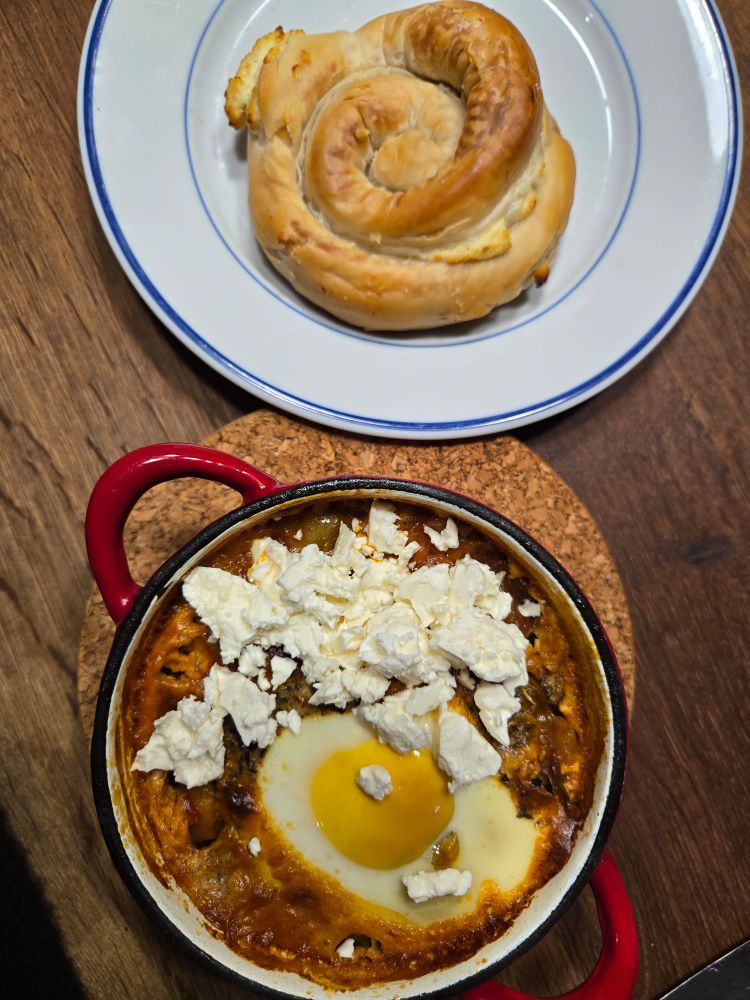 A red cast iron full of tomato sauced giant beans topped with and egg and feta crumbles and a phyllo pastry dough based spiral on an adjacent plate. 