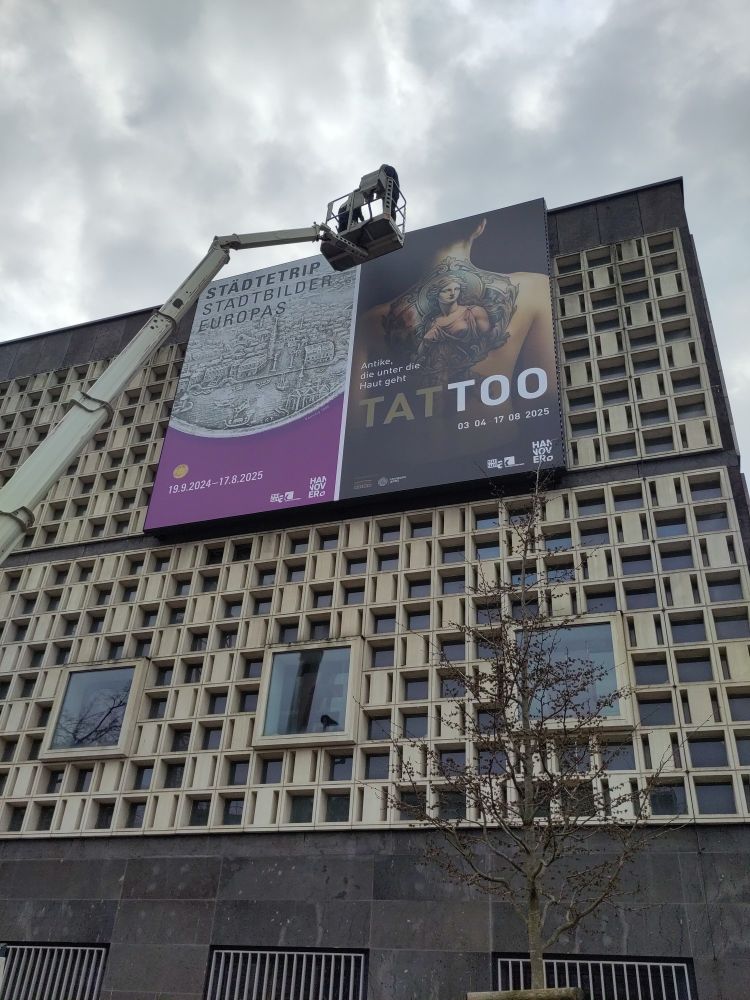 The facade of Museum August Kestner (Hannover, Germany), a modern building with a geometric square pattern all over it. At the top hang two banners: The left one shows a silver medallion of magenta ground and the text "Städtetrip. Stadtbilder Europas" and "19.9.2024 - 17.8.2025".
The left one shows the naked back of a woman with a large tattoo of a woman in a classicist style. The background is black. The text reads "Tattoo. Antike, die unter die Haut geht. 03.04. - 17.08.2025"

A crane is hovering in front of the banners.