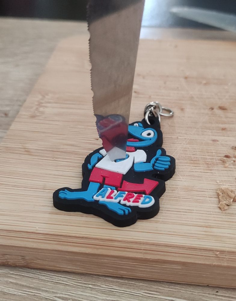 Foto von einem Echsen-Schlüsselanhänger. Die Echse ist blau und trägt ein weiszes T-Shirt mit blauem Herz, und eine kurze rote Hose. Der Schwanz der Echse ist ein roter, nach oben gebogener Pfeil. Der Name der Echse ist auch am Fuszende vermerkt,Alfred. Hier sind die Buchstaben eins, drei und sechs in blau, die anderen in rot.
Das blaue Herz auf dem Shirt wird von einer Messerspitze aufgespieszt. Der Anhänger liegt auf einem Holzbrett.