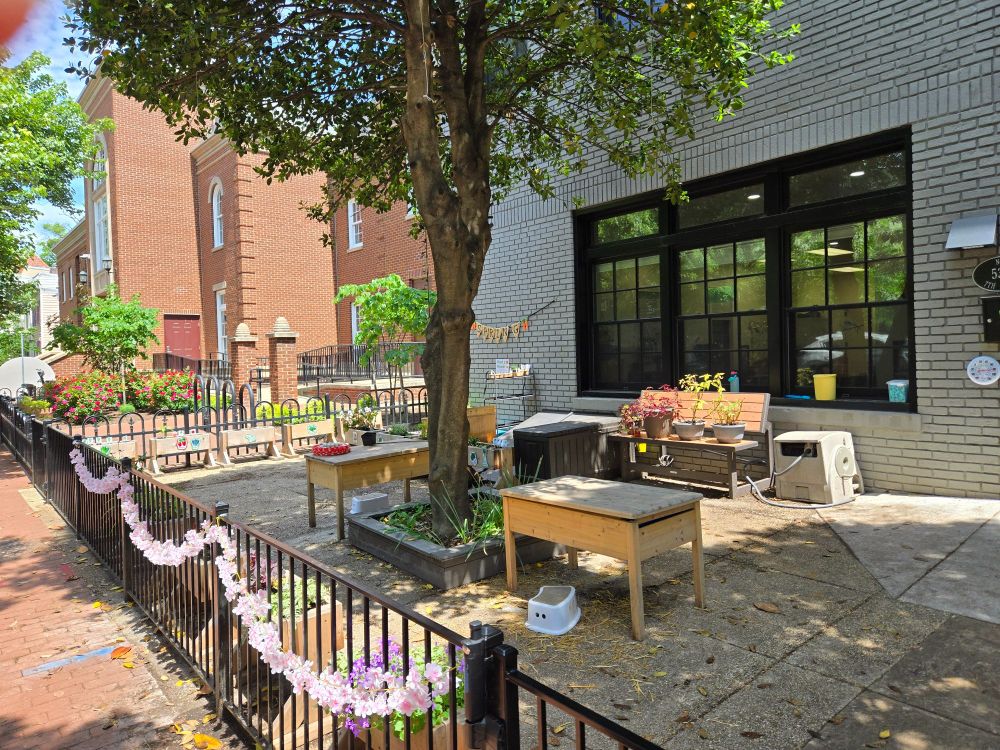 The patio of a Montessori preschool with several mini planters full of plants, benches, and stools for the kids. A tree is in the middle & the fences are hung with decorations. 