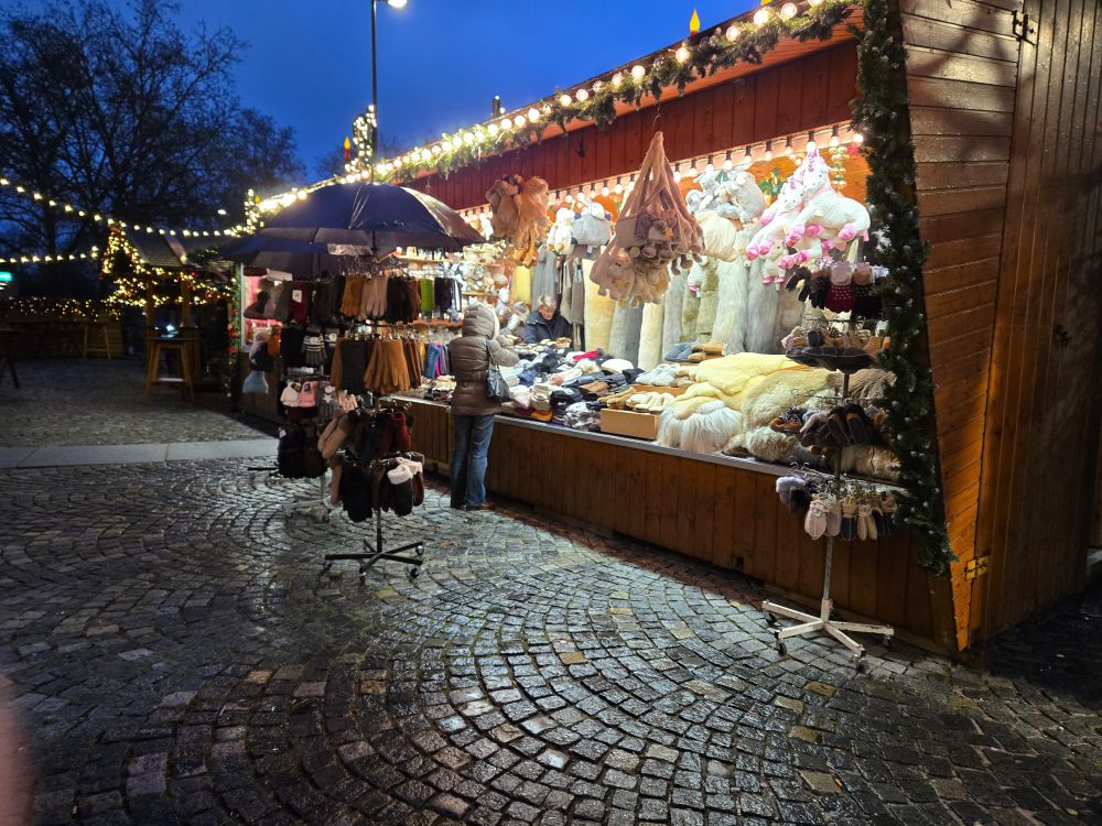 A well-lit miniature wooden lodge decorated with festive greenery & displaying a variety of hats,  gloves, scarves,  and other hand made fabric goods. 