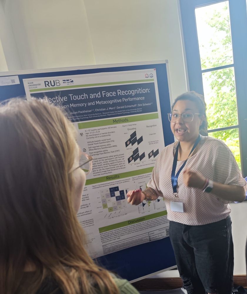 a mixed race person wearing a black pair of pants, a pink blonde and a blue lanyard standing in front of a scientific poster titled „affektive touch and face recognition“. they are in the middle of wxplaining something, however they noticed the camera person and looked at the camera 