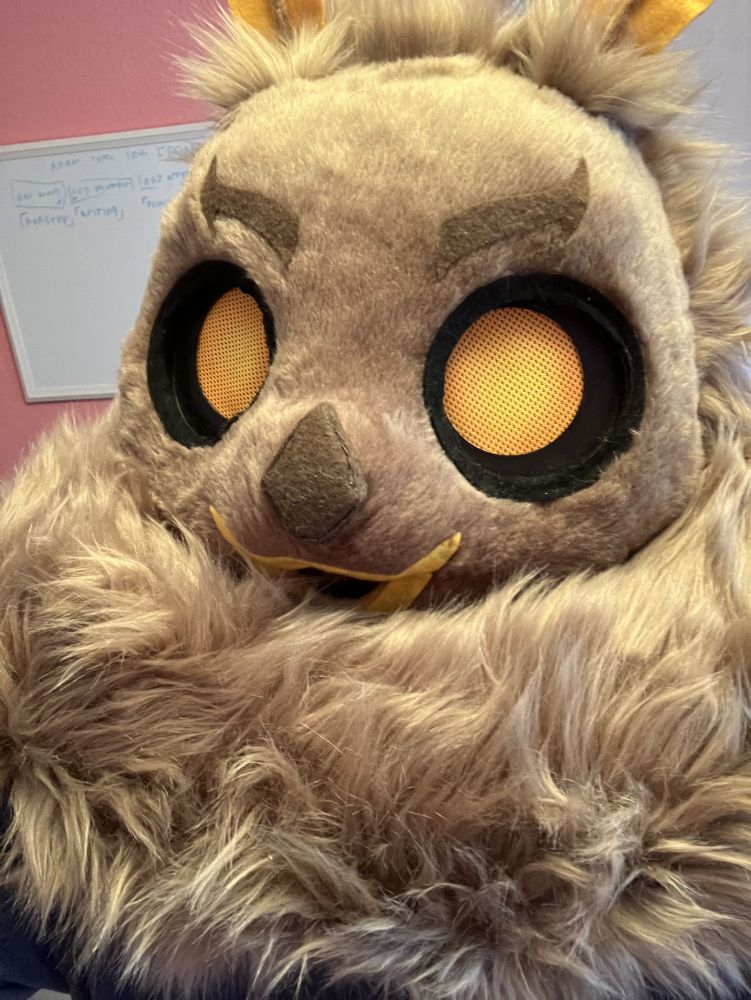 A brown moth fursuit head with orange eyes looking at the camera with a large mane of neck fluff below