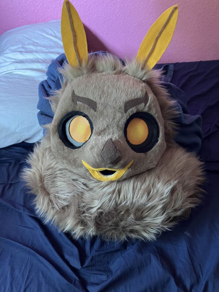 A brown moth fursuit head with orange eyes looking at the camera with a large mane of neck fluff below