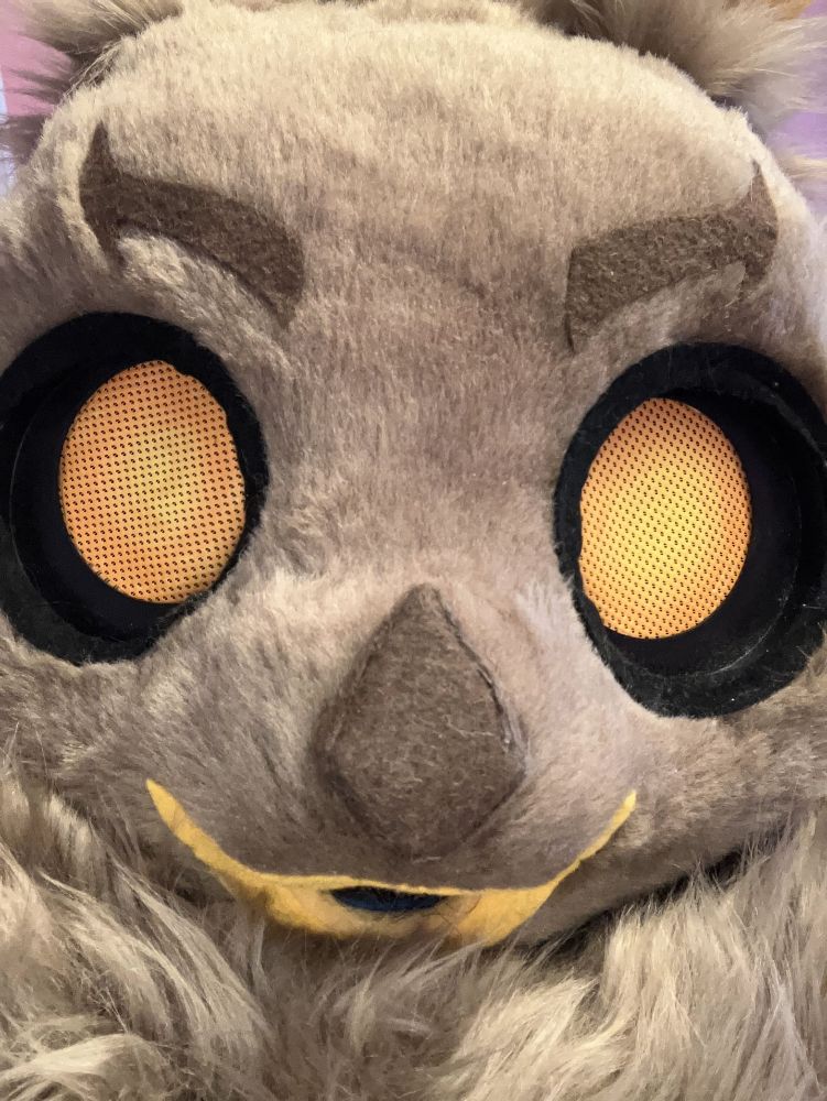 A brown moth fursuit with orange eyes looking at the camera with a large mane of neck fluff below