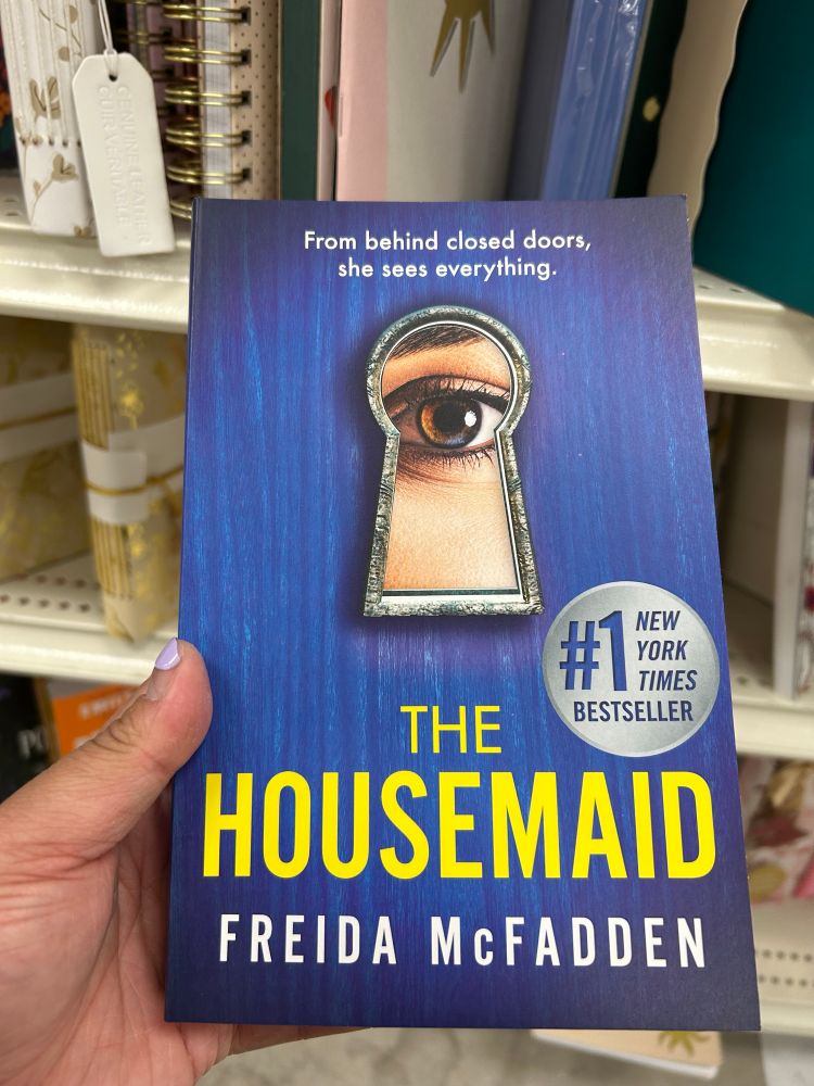 Holding a paperback novel with my left hand. A blue cover with an eye peeking through a lock hole. The title reads “The Housemaid” with subtitle “Freida McFadden” (author) in block letters. Also a silver badge on the right reads “Number 1 New York Times Bestseller”. 