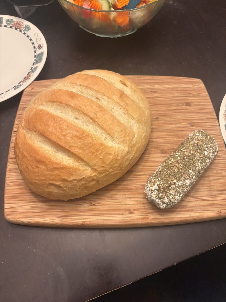 A large loaf of bread and a herb-covered chèvre on a wooden board. 