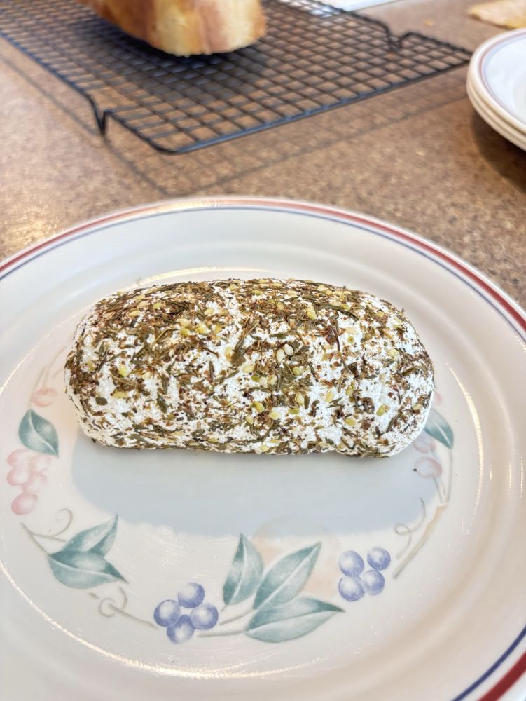 A log-shaped roll of chèvre covered with herbs on a plate with grapes-and-leaves design. 