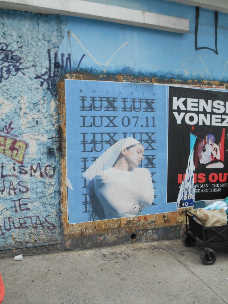 foto de um muro azul claro com pixos e cartazes. no meio há um cartaz azul claro com a divulgação do disco lux em 07.11 e a cantora rosalía vestida de freira, em branco, com os olhos fechados olhando para o lado