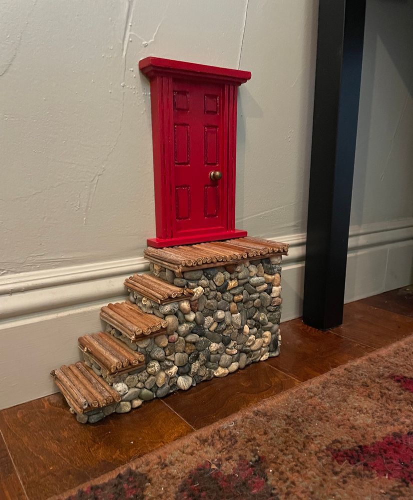 Red Fairy door above the baseboard of our entryway, set upon a staircase made of blackberry bush stems and gravel from the garden.