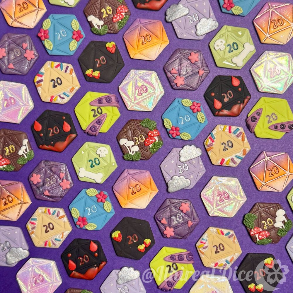 Many flat polymer clay d20 charms of different designs laying on a purple background. 