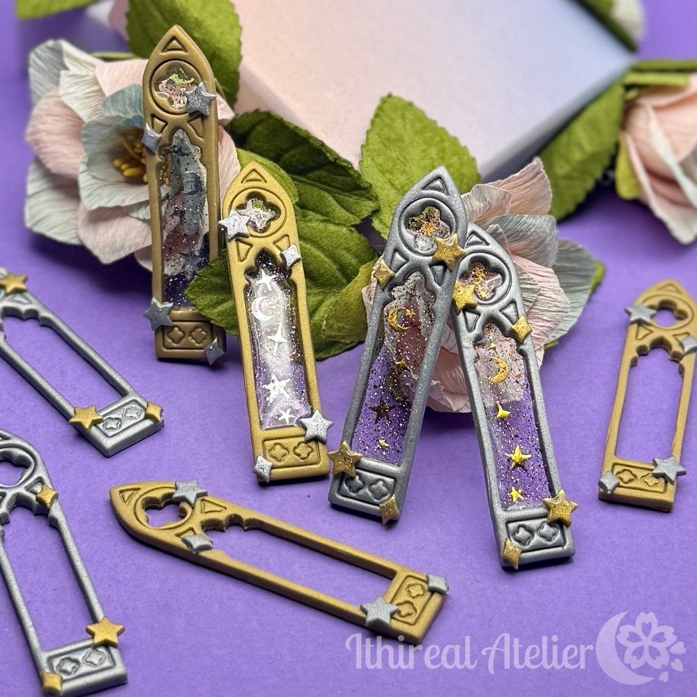 A pair of gold and silver polymer clay cathedral windows with glittery transparent “glass” with stars and moons inside, surrounded by empty window frames and resting on a fake flower. 