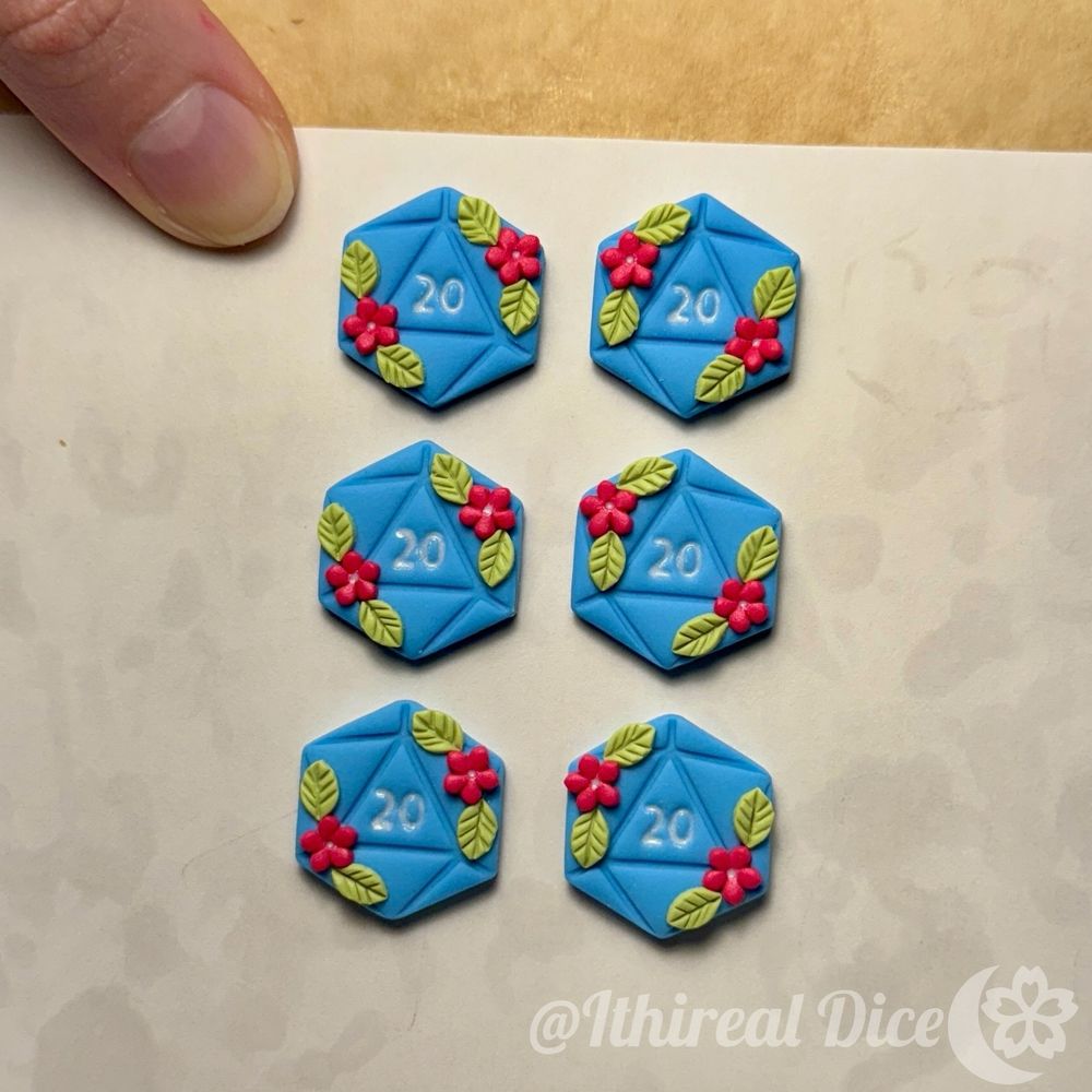 Six unbaked blue polymer clay d20 earring charms. They have bright pink flowers with green leaves on them. The 20 in the middle is white. They are on a baking sheet, held by my thumb. 