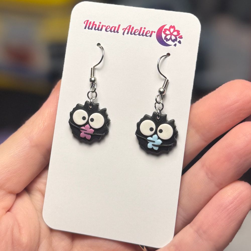A pair of polymer clay earrings that look like soot sprites holding purple and blue konpeito, on a white backing card that says Ithireal Atelier, in my hand. 