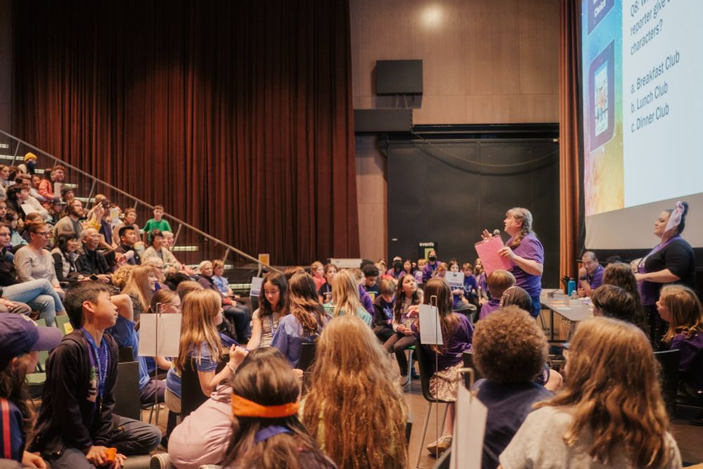 A librarian speaks to children in an auditorium.
