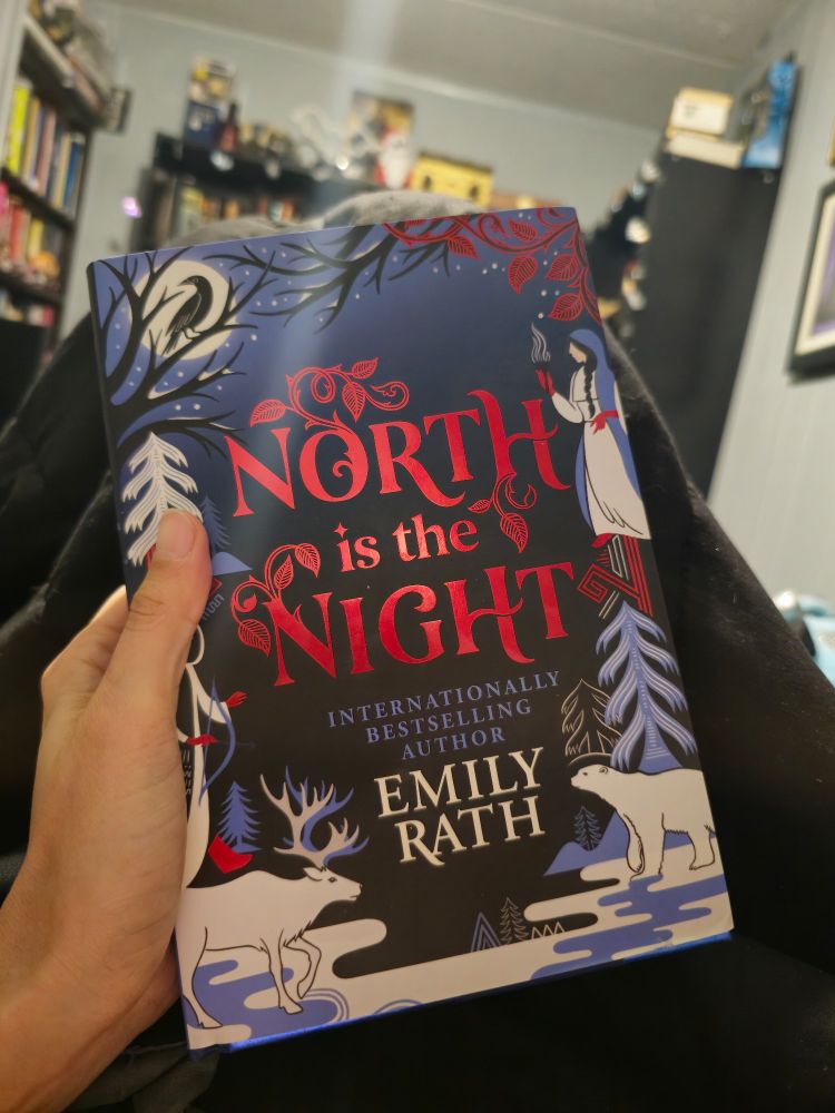 My hand holding a hardcover copy of North is the Night by Emily Rath the title is in red and author name in white. Dark blue cover with blue and white artwork along the edges featuring a reindeer, polar bear, a dark haired girl, birds and various trees and snow.