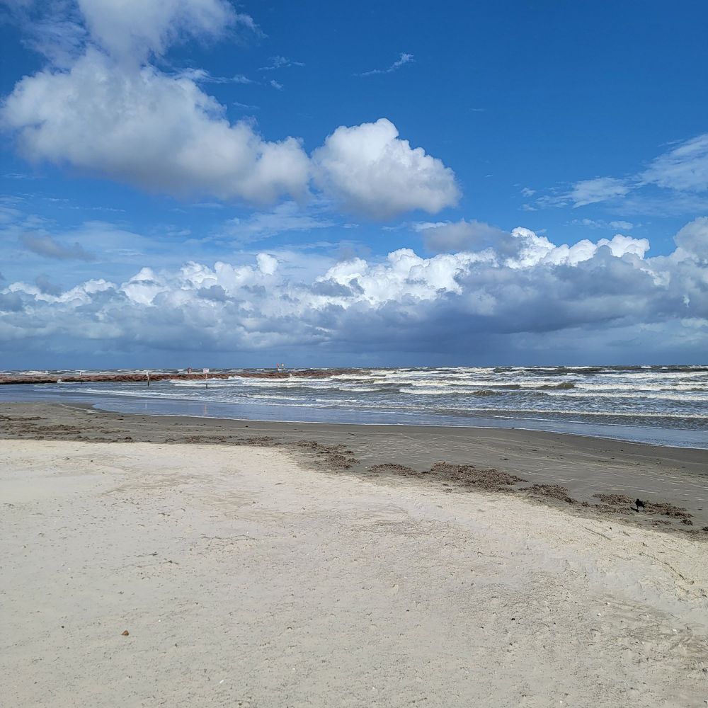 Galveston Island, Gulf of Mexico. Empty beach with murky water full of waves and a blue sky with white clouds in the forefront and dark storm clouds in the the distance