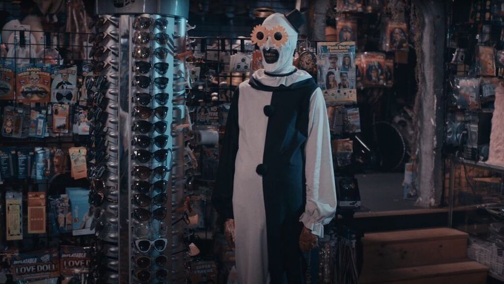 A still of Art the clown from terrifier 2 standing in a halloween store next to a sunglasses rack wearing giant sunflower sunglasses with a sinister smile on his face while standing straight with his arms to his side. He is wearing white face paint with his mouth painted black and has on a black and white clown suit.