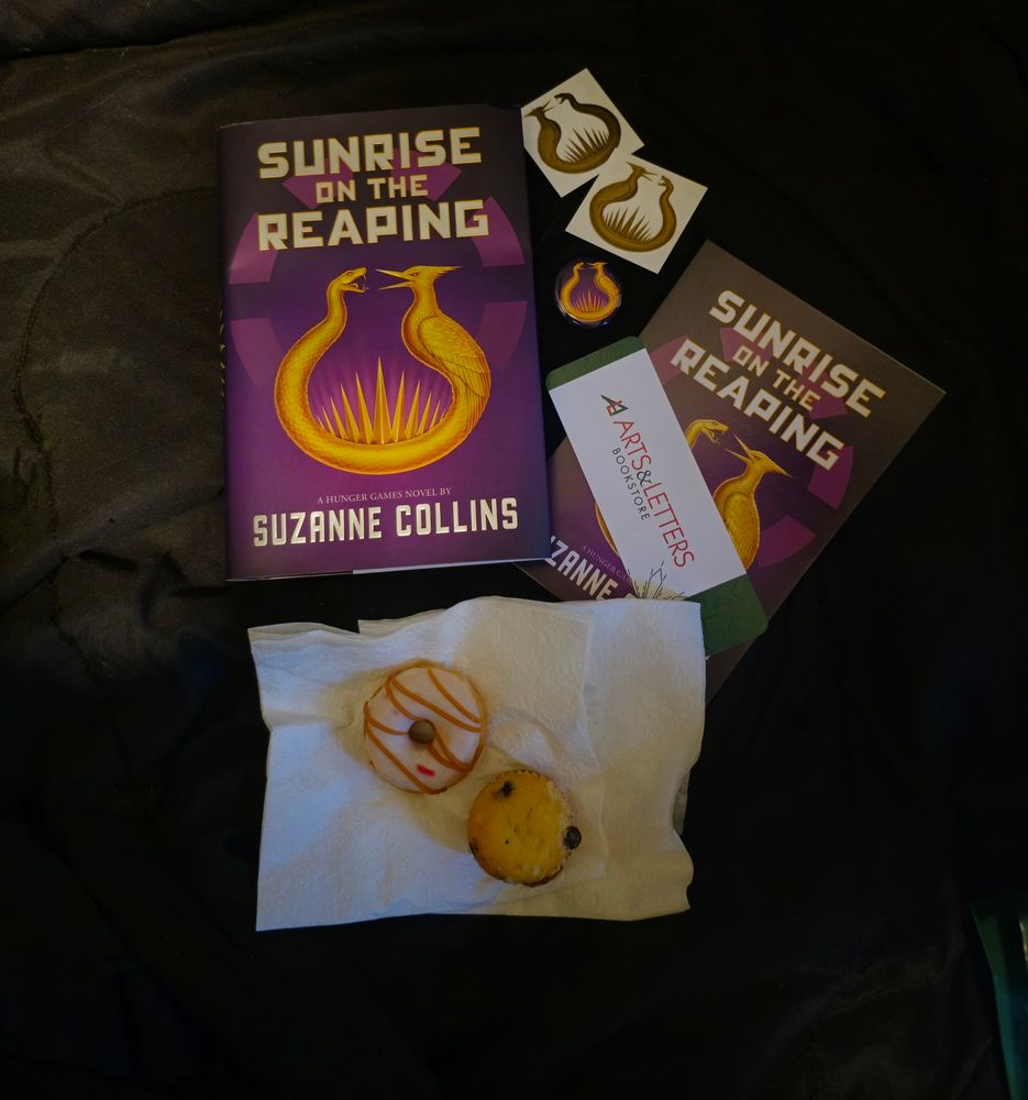 A hardcover copy of Suzanne Collins' Sunrise on the Reaping sitting beside 2 temporary tattoos that matches the gold snake/mockingjay emblem on the book, a purple and gold pin that has the same emblem as the book, a postcard featuring the book's cover, a red white and green bookmark that says "Arts & Letters Bookstore, and a mini donut with white icing and caramel drizzle and a blueberry mini muffin sitting on top of a white paper napkin