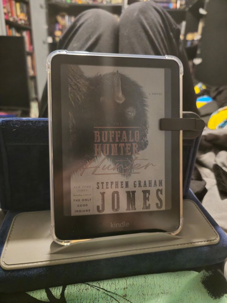 Kindle Colorsoft sitting on a blue pillow tablet stand with the cover of The Buffalo Hunter Hunter displayed on the screen.