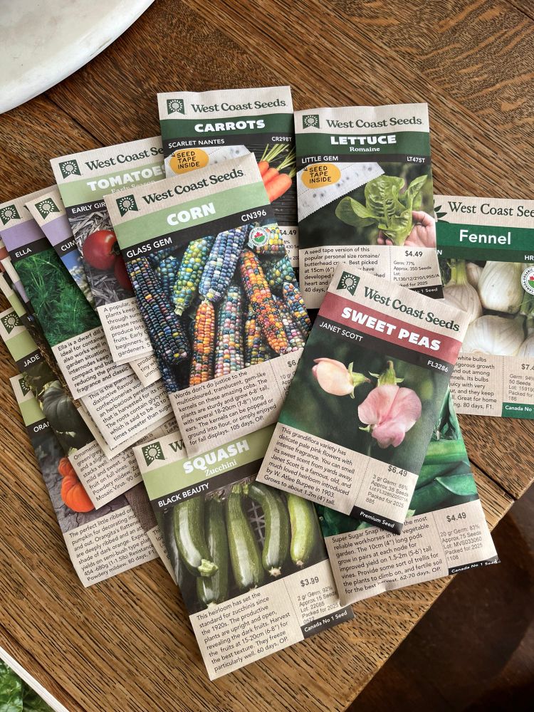 Many packets of seeds from West Coast Seeds are spread on a wooden table; some visible ones include sweet peas, glass gem corn, fennel, and lettuce.