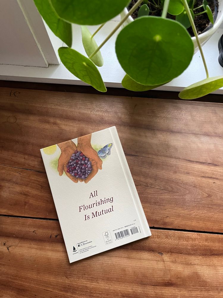 A photo of the back of Robin Wall Kimmerer’s book “The Serviceberry” with an illustration of hands cupped, holding berries and the words “All Flourishing is Mutual”. It is on a wooden table with a few green pilea leaves peeking in.