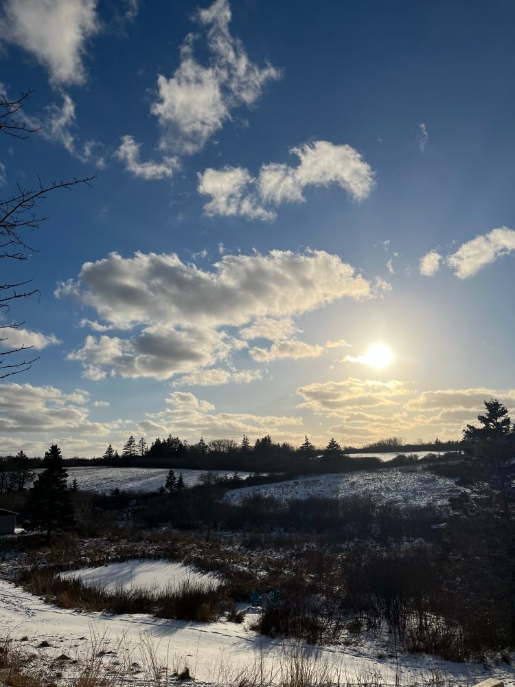Sun peeks through a sky of clouds over snow-covered fields and a pond.