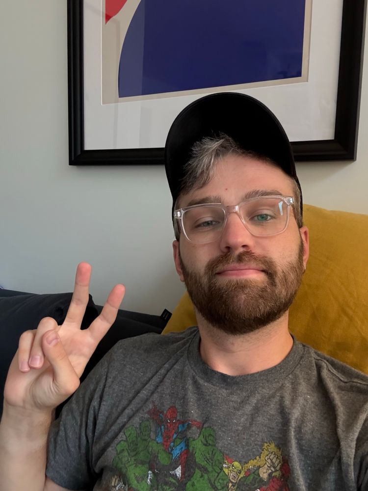 Kyle - wearing a hat and a grey pajama shirt, putting a peace sign up.