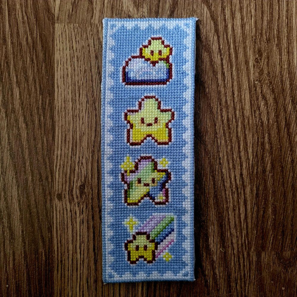 Marque-page brodé avec des motifs de petites étoiles souriantes sur fond bleu, posé sur un sol en bois.