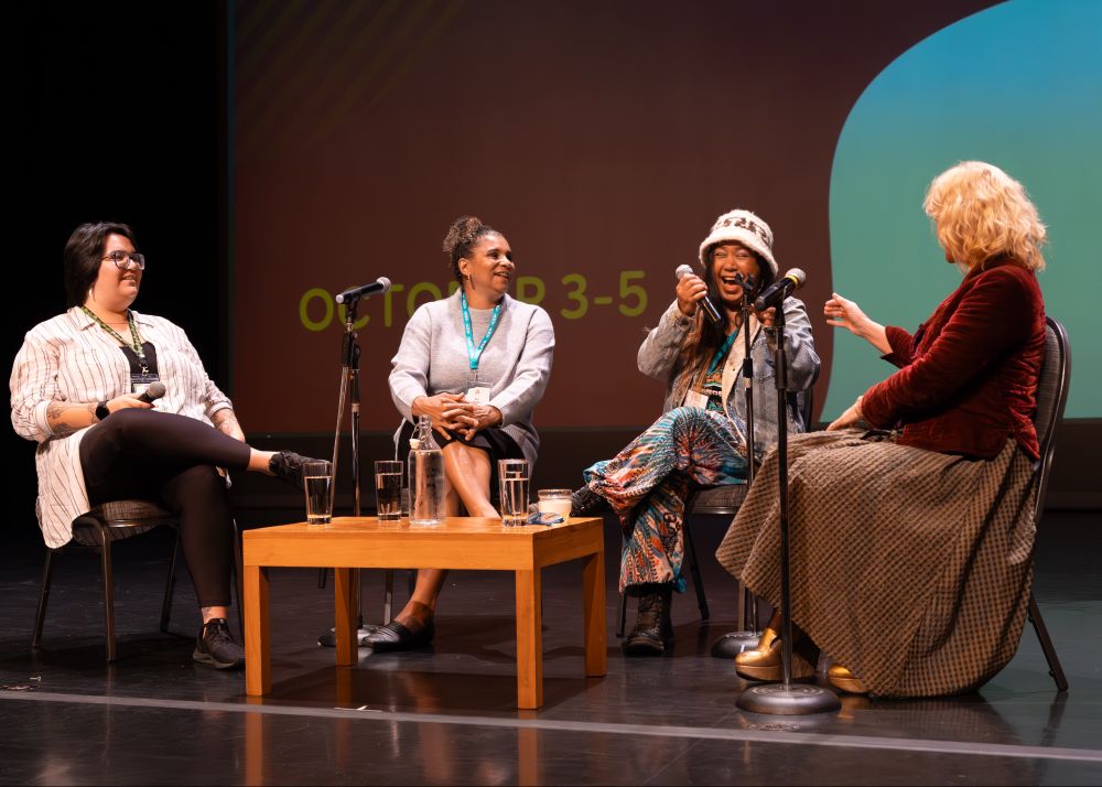 Four panelists sit in a semi-circle on a stage, engaged in a lively conversation during a conference. They are smiling and laughing, with microphones in front of them. The setting appears warm and collegial, suggesting a thoughtful and joyful discussion. Photo by Eri Cardenas
