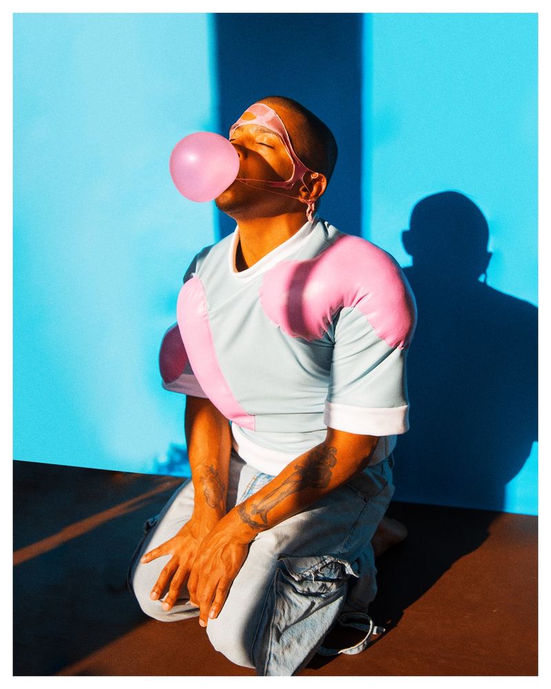 full body self portrait blowing a bubble with bubblegum on my knees. a previous is bubble is popped all over my face. gum is  bright pink against a blue backdrop. bubbles are expanding out of the baby blue shirt i am wearing.