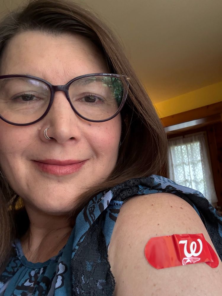 A woman wearing glasses smiles and shows her bare upper arm, which has a red Walgreens bandaid on it