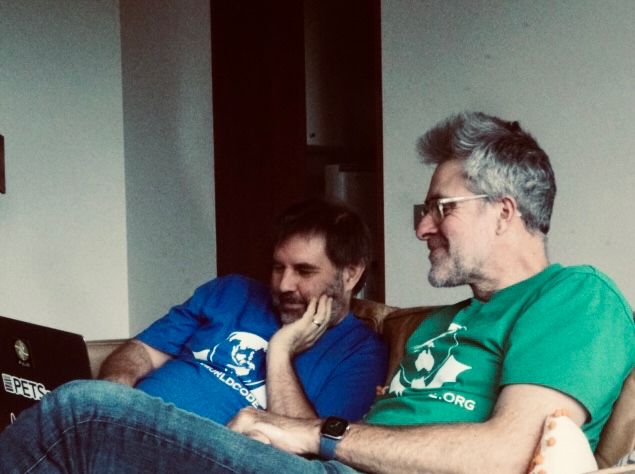 Thing one and thing two sitting on a sofa working on laptops. Classic human pretending to ponder deeply poses (scratching chin / biting nails). The T shirts match but one is blue and two is green. It may be the other way around.