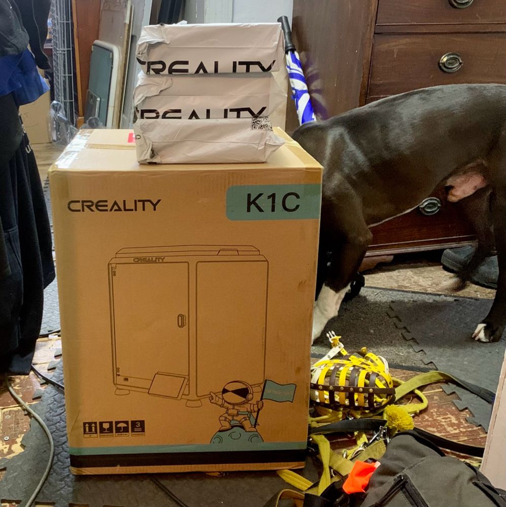 A photo of a Creality K1C 3D printer box and three parcels containing printing filament that had just been dropped off by courier. A medium sized brown and white dog (~40kg) is partially obscured by the box, as he’s having a good sniff at the side away from the camera. His bright yellow harness, lead, and muzzle are being partially squished by the box, as it was heavy and I kinda put it down in a hurry when the courier dropped it off. The rest of the hallway, as my house (and arguably life), is a mess, so I’ll not dwell on describing that in too much detail. Composition is terrible, but I’m no David Bailey. Taken on an old iPhone, with a level of quality appropriate for hardware of the time. 