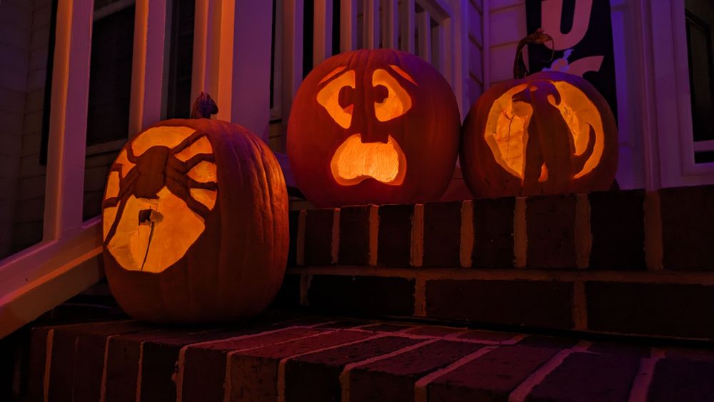 Three Jack-o'-lanterns on a front porch. One is a spider, one is a goofy face and one is a dog in profile.