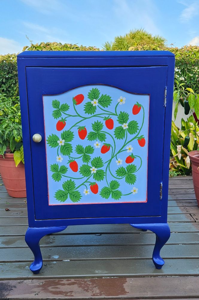 A little bright blue bedside cabinet with a panel of lighter blue on the door painted with a sinuous pattern of strawberry vines and flowers