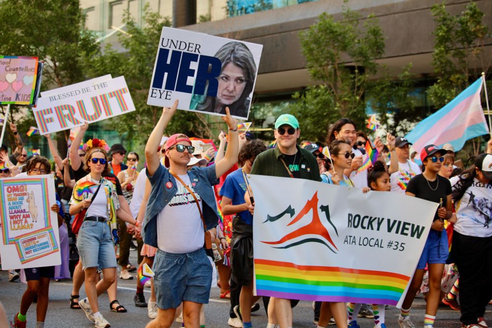 Photo of a person wearing a red baseball cap backwards, sunglasses, blue shorts and a t-shirt with the words, “Protect Trans Kids” while holding up a sign showing Danielle Smith with a scowling face and the words, “Under HER eye.” Next to them is another person with a bright blue baseball cap, dark sunglasses and holding a banner with the words, “Rocky View ATA Local #35” and a rainbow, with a smile for the camera. Other signs held by many other people in the background have the words, “Blessed be the FRUIT,” “School counsellors protecting kids,” and “OMG! My teacher read me a book about llamas…Why am I not a llama, Learning about LGBTQ is understanding not becoming.” There are many rainbow flags and a trans flag. 