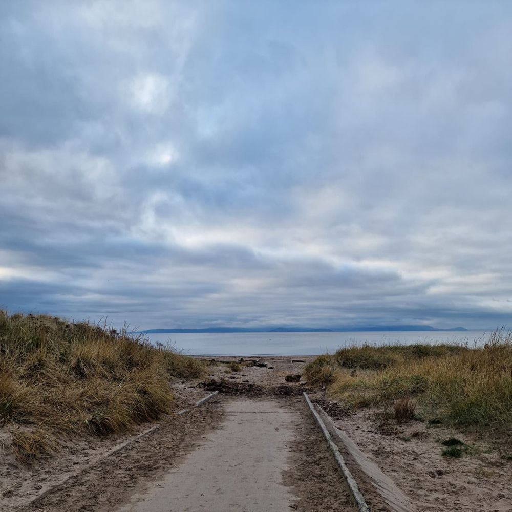 A concrete path, with sand all along the edges, stretching out through grassy dunes to a sandy beach, the sea beyond and a low, dark, island on the horizon. The sky is full of interesting clouds