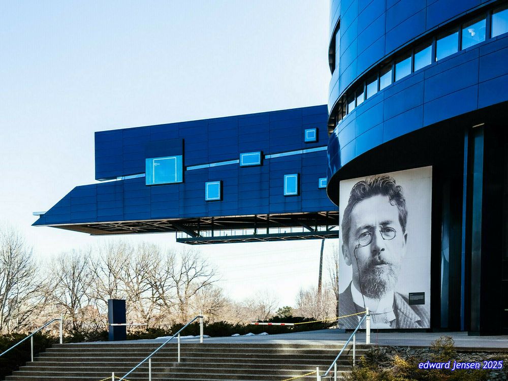 Modern blue architectural building with a distinctive cantilevered section featuring glowing blue windows. A large black and white portrait of a bearded man with glasses is displayed on the exterior wall. The building has concrete steps with metal railings leading up to the entrance, set against a backdrop of bare winter trees and clear sky.