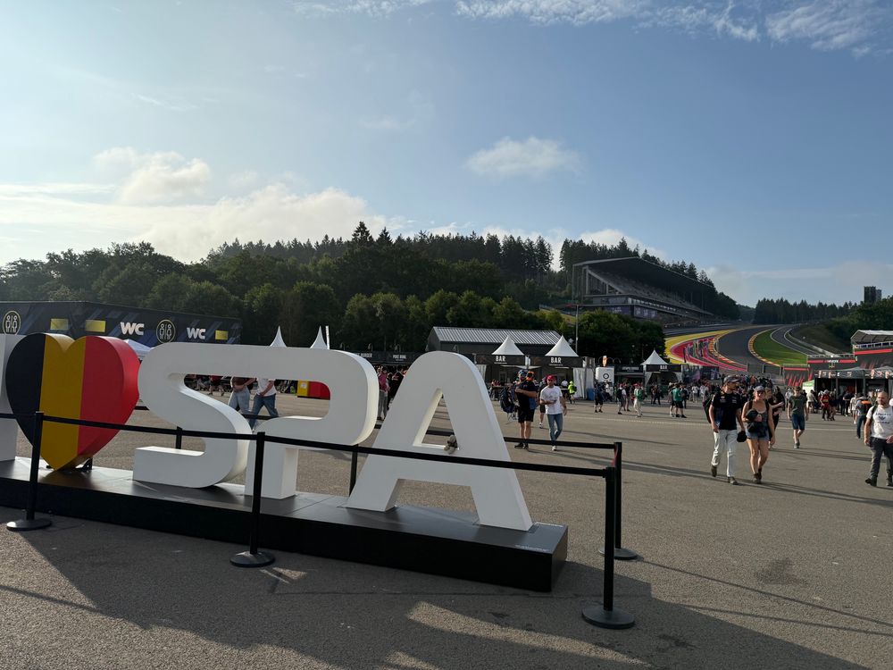 View of the fanzone area at Spa-Francorchamps, with Eau Rouge and Raidillon visible behind. There’s a sign in the foreground that says ❤️Spa (with the heart in the colours of 🇧🇪).