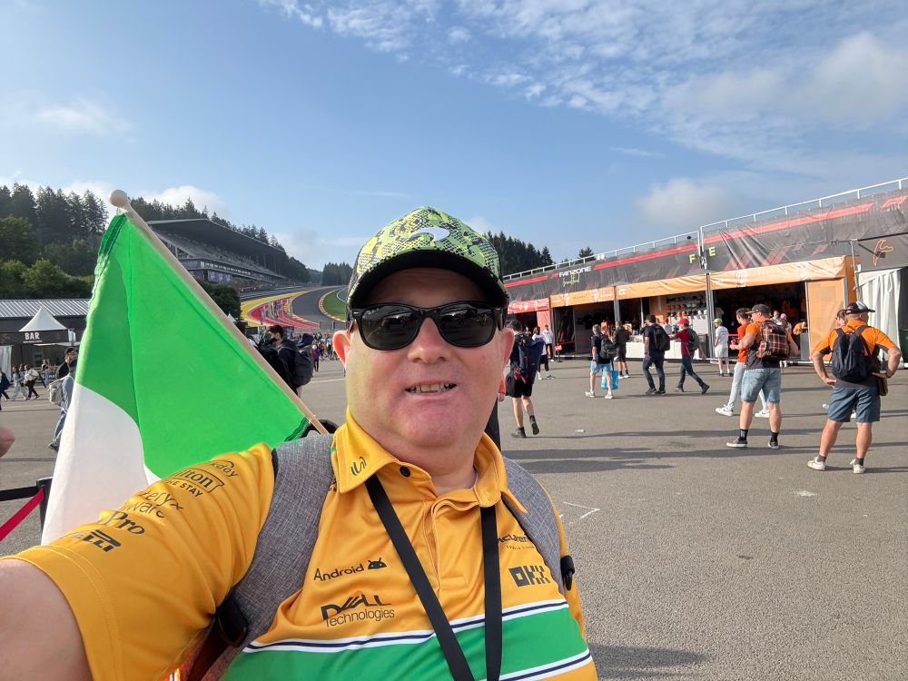 Paul standing in the Fanzone at Spa-Francorchamps wearing a yellow and green Senna-McLaren top, a green McLaren cap and sunglasses. A 🇮🇪 is sticking out of his backpack.