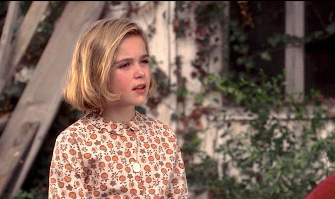 Screenshot from the tv show Mad Men of Sally Draper, a blond girl, talking. She is sitting outside and you can see a house with greenery in the background. Salt has a blond Bob and is wearing a cream shirt with orange pumpkins? printed on it.