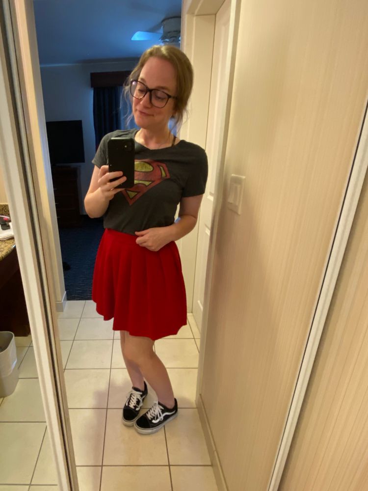Mirror selfie of Megan, a brunette, wearing a grey superman tshirt with a pleated red skirt, black vans, and black “Clark Kent”-style glasses 