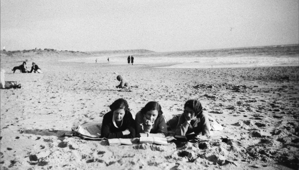 Three women lying on a beach reading.