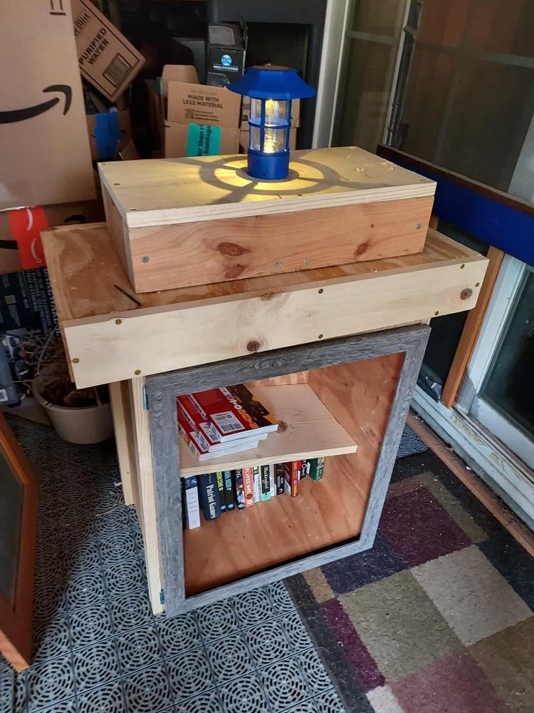 A wooden little free library, that vaguely resembles the TARDIS with a box on top. It has books inside, including a shelf on top of them.