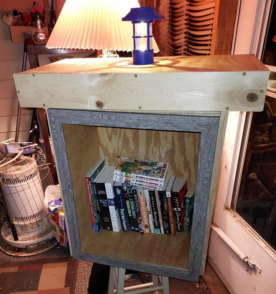 My little free library with the box attached on top, a wooden frame for the door on the front, and the books inside. There is a lit lantern on top.