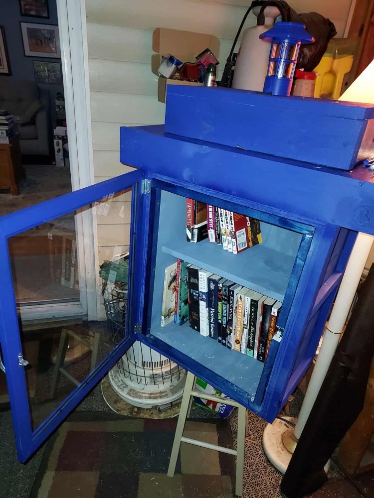 The same little free library with the door open, showing a reflection in the glass, a dark blue frame on the inside of the door, and the books inside.