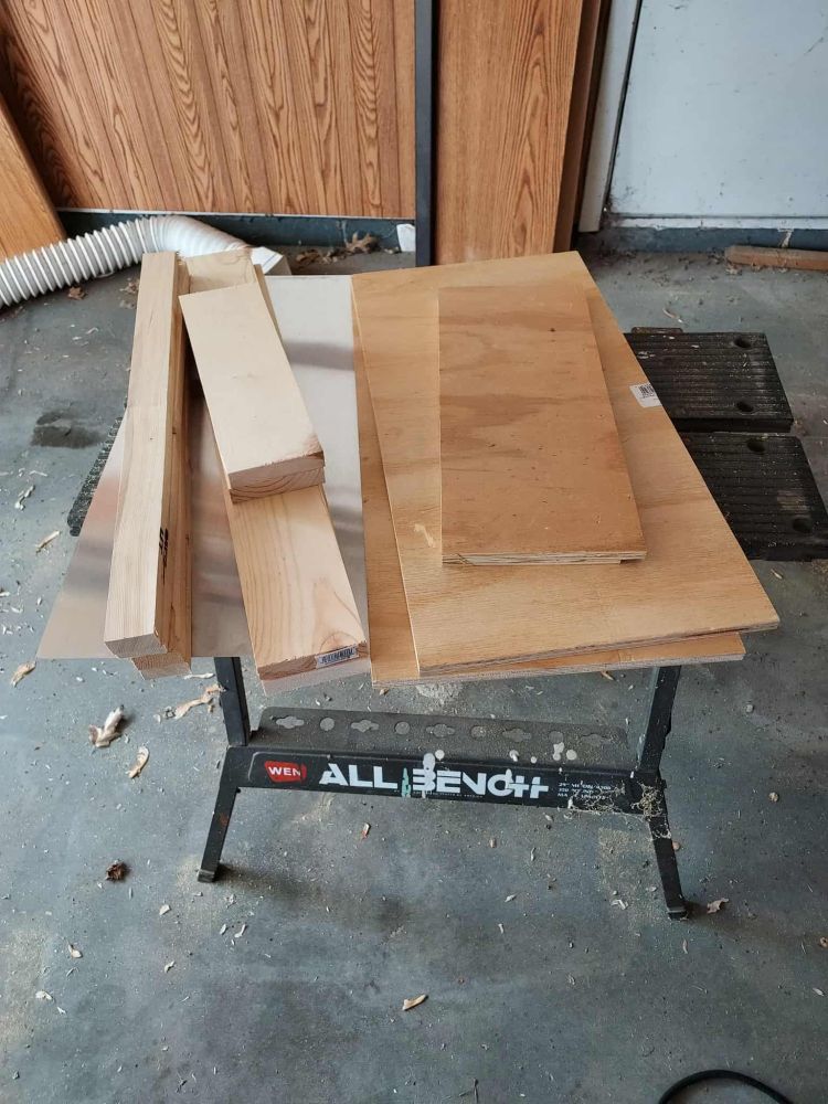 Some dress-up bits for my little free Library, cut lumber sitting on a work bench. IT's also sitting on a sheet of aluminum that will be a sort of rain shield for the top of it.